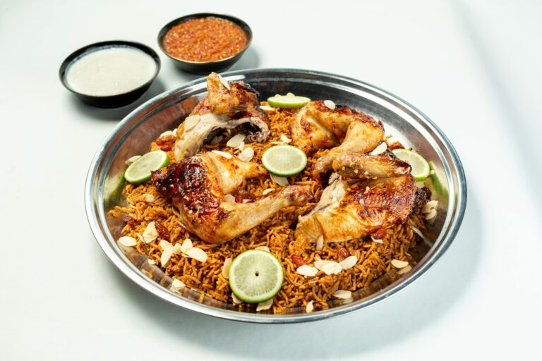Close-up of chicken biryani with slices of lime, almonds, and sauces on a white background.